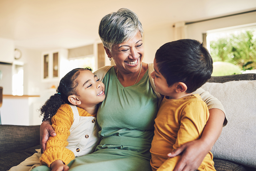 older woman smiling and hugging a her granddaughter and grandson on the couch in her home while they smile back gold shirts